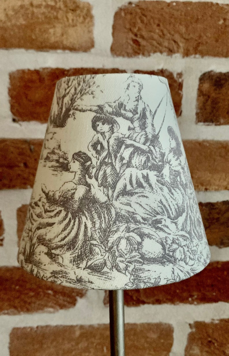 May include: A small, off-white lampshade with a detailed gray toile design featuring figures in a pastoral scene. The lampshade is cone-shaped and mounted on a silver metal stand. The background is a brick wall.