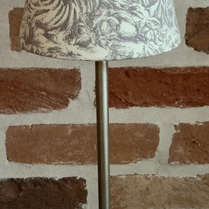 May include: A decorative table lamp with a silver-colored base and stem. The lampshade features a cream-colored background with a gray toile design depicting figures in a pastoral scene.
