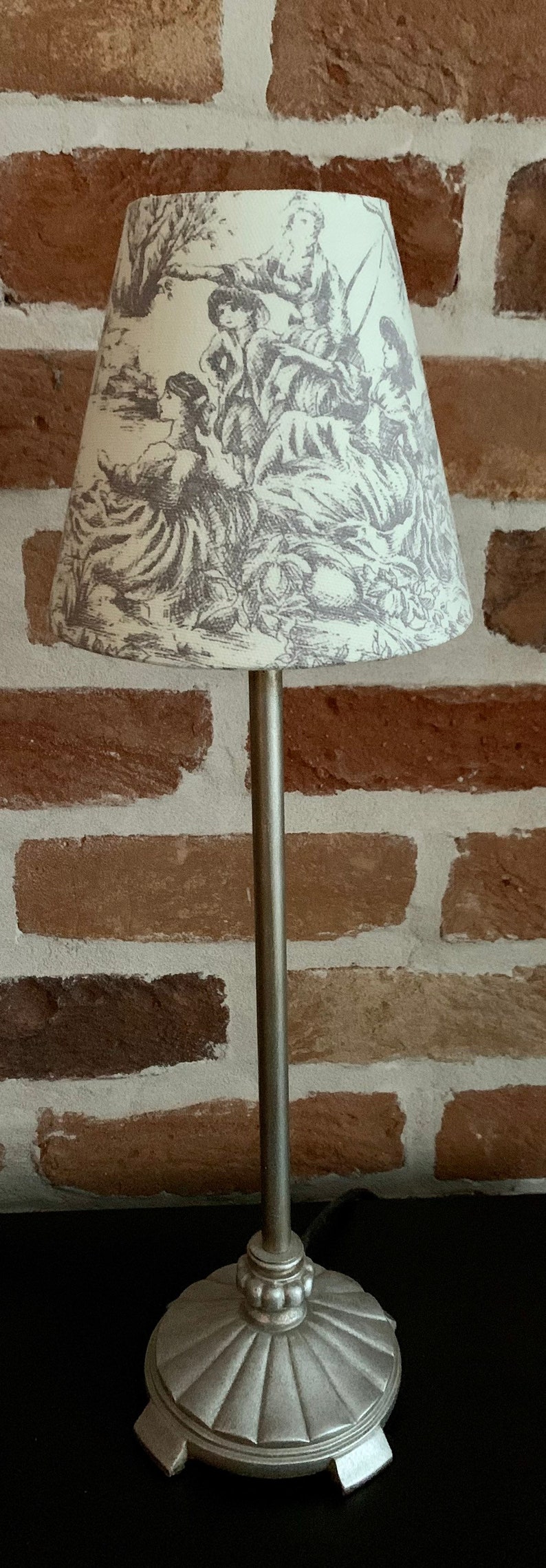 May include: A decorative table lamp with a silver-colored base and stem. The lampshade features a cream-colored background with a gray toile design depicting figures in a pastoral scene.
