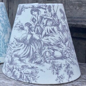 May include: A white lampshade with a gray toile pattern depicting figures in a pastoral scene. The lampshade has a conical shape and is made of fabric. The design features trees, flowers, and people in period clothing.