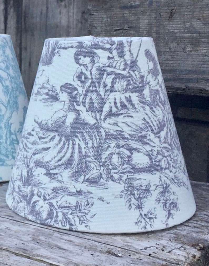 May include: A white lampshade with a gray toile pattern depicting figures in a pastoral scene. The lampshade has a conical shape and is made of fabric. The design features trees, flowers, and people in period clothing.