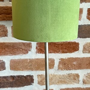 May include: A table lamp with a light green lampshade and a silver-coloured base. The lampshade is cylindrical and made of a soft, textured material. The base has a decorative, ornate design. The lamp is on a black surface.