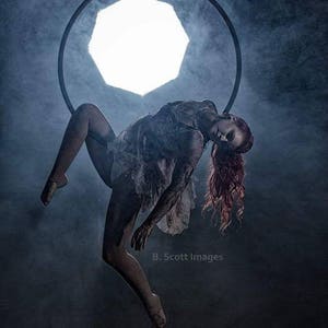 May include: A woman with long red hair hangs upside down in a hoop, suspended from the ceiling. The hoop is made of metal and is circular. The woman is wearing a white dress and has a smoky background.