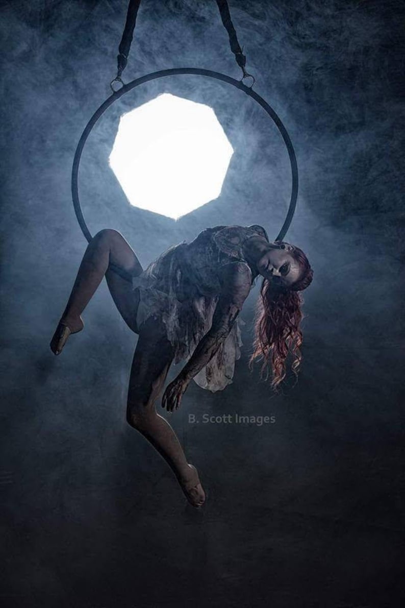 May include: A woman with long red hair hangs upside down in a hoop, suspended from the ceiling. The hoop is made of metal and is circular. The woman is wearing a white dress and has a smoky background.