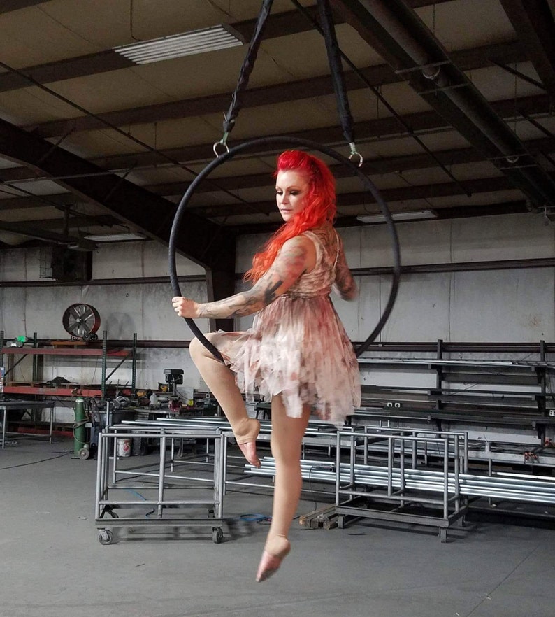 May include: A woman with red hair performs an aerial hoop routine in a warehouse setting. She is wearing a light brown dress and is suspended in the air by a black hoop.