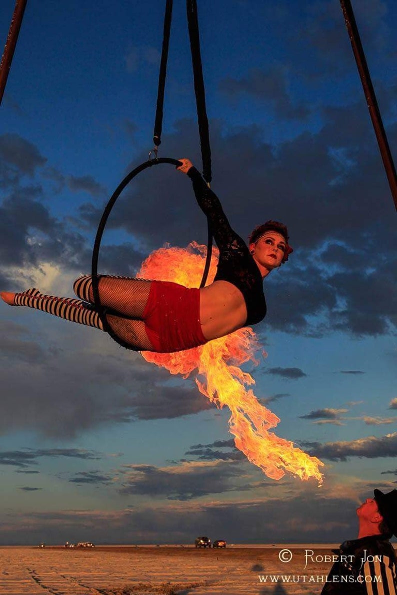 May include: A woman performs an aerial hoop routine with fire emanating from the hoop. She is wearing a red and black outfit and is suspended in the air against a blue sky with clouds. The image is taken at Burning Man.