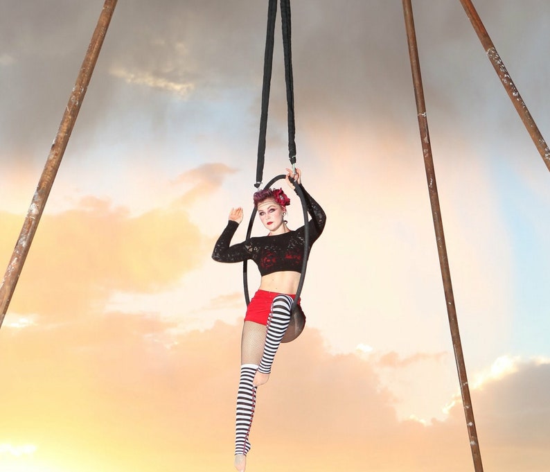 May include: A woman in a black and white striped outfit performs an aerial hoop routine. She is suspended from a hoop hanging from the ceiling. The background is a cloudy sky.