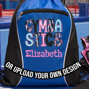 May include: A blue and black drawstring gym bag with the word "GYMNASTICS" in colorful letters and the name "Elizabeth". A purple water bottle, gym chalk, and gymnastics apparel are also visible.