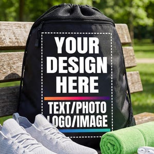Op de afbeelding: Een zwarte sporttas met trekkoord met de tekst "YOUR DESIGN HERE TEXT/PHOTO LOGO/IMAGE" wordt getoond op een houten bank. Een paar grijze sportschoenen, een opgerolde groene handdoek en witte oortelefoons maken de scène compleet.