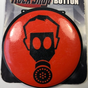 May include: A red round button with a black gas mask graphic. The text "Rock Shop Button" is printed on the background.
