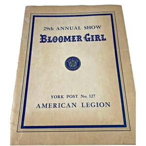May include: A tan paper program cover for the 29th Annual Show Bloomer Girl, featuring a blue border, blue text, and a central American Legion emblem. The text reads 29th ANNUAL SHOW BLOOMER GIRL, YORK POST No. 127, and AMERICAN LEGION.