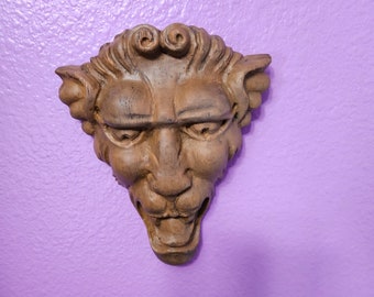 May include: A detailed, brown, carved wooden face with a stern expression, mounted on a purple wall. The face has a prominent nose, furrowed brow, and curly hair. The sculpture is likely a decorative wall hanging.