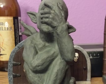 Può includere: Una statua di gargoyle grigia con le ali ripiegate, che si copre gli occhi con le mani. La statua è seduta con le gambe incrociate e i piedi visibili. La statua è su una superficie di legno.
