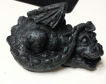 Puede incluir: Una pequeña figura de dragón durmiendo hecha de piedra gris oscura. El dragón tiene una cola enroscada, los ojos cerrados y una pequeña boca abierta.