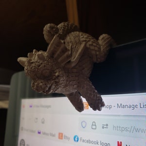 May include: A gray ceramic dragon figurine with detailed scales and wings. The dragon is perched on the edge of a computer screen, with the text "Shop - Manage List" visible in the background.
