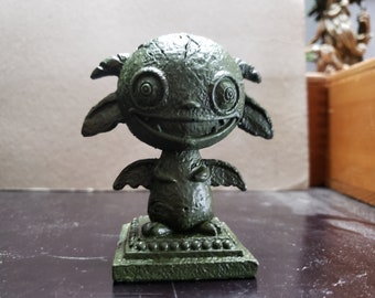 Può includere: Una statuetta verde, dettagliata, stampata in 3D di una piccola creatura alata con un ampio sorriso e grandi occhi rotondi. La creatura è in piedi su una base quadrata con un bordo decorativo.