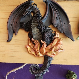 Puede incluir: Una figura de drag&oacute;n negro con las alas extendidas, posada sobre una base de llama dorada. El drag&oacute;n tiene una textura escamosa detallada y una expresi&oacute;n feroz.