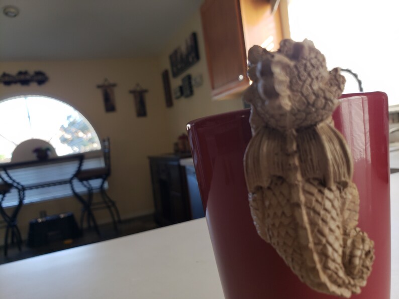 Puede incluir: Una taza de cer&aacute;mica roja con una figurita de drag&oacute;n marr&oacute;n unida al lado. El drag&oacute;n est&aacute; mirando la taza y tiene un cuerpo texturizado y escamoso.