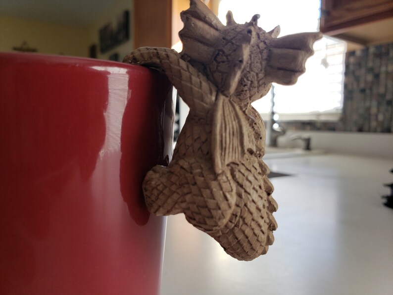 Puede incluir: Una figurita de drag&oacute;n de cer&aacute;mica con una superficie texturizada, trepando por el lado de una taza roja.