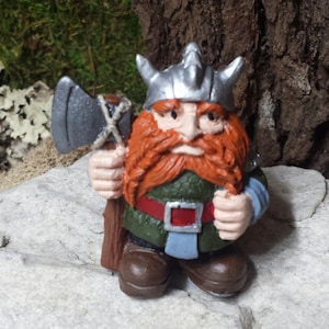 May include: A small, hand-painted figurine of a Viking warrior with a red beard, wearing a silver helmet and holding an axe. The figurine is standing on a gray rock.