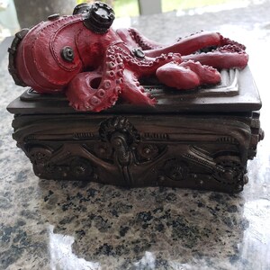 K&ouml;nnte beinhalten: Eine dunkelbraune, kunstvoll gestaltete Box aus Metall, auf der ein roter Oktopus mit Steampunk-Details thront. Die Box hat einen Klappdeckel.