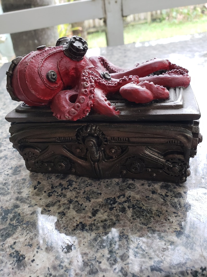 K&ouml;nnte beinhalten: Eine dunkelbraune, kunstvoll gestaltete Box aus Metall, auf der ein roter Oktopus mit Steampunk-Details thront. Die Box hat einen Klappdeckel.