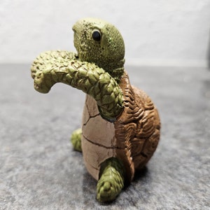 May include: A ceramic turtle figurine with a detailed, textured shell in shades of brown and green. The turtle's head and limbs are green, while the shell is brown with visible cracks. The figurine is posed as if it is crawling.