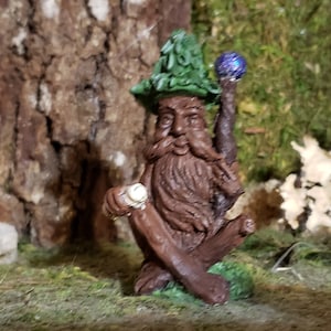 May include: A small, brown, wooden gnome figurine wearing a green hat and holding a staff with a blue orb on top. The gnome is sitting on a tree stump and has a long, white beard.