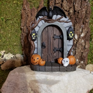 May include: A miniature Halloween door with a bat above it, a wooden door with black iron accents, and two lit lanterns on either side. There are three pumpkins and a skull on the ground in front of the door.