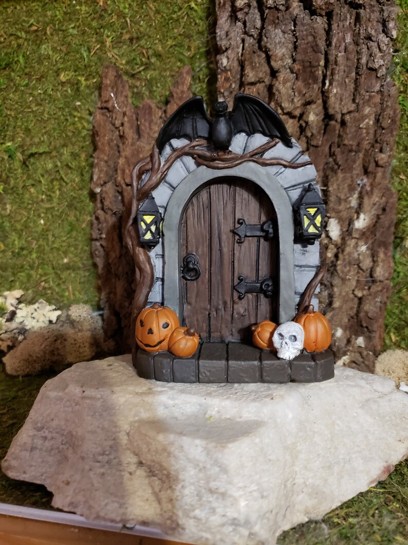 May include: A miniature Halloween door with a bat above it, a wooden door with black iron accents, and two lit lanterns on either side. There are three pumpkins and a skull on the ground in front of the door.