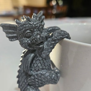 Puede incluir: Una peque&ntilde;a figura de drag&oacute;n gris con escamas y alas texturizadas. El drag&oacute;n se aferra al lado de una taza de cer&aacute;mica blanca.