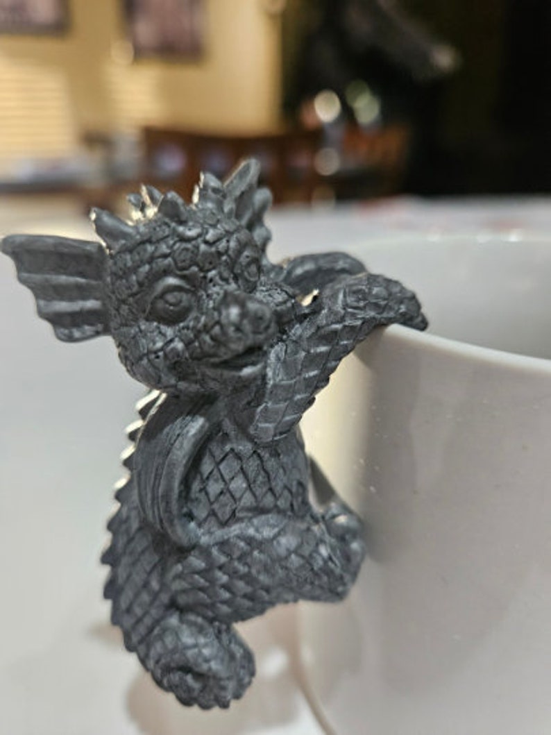Puede incluir: Una peque&ntilde;a figura de drag&oacute;n gris con escamas y alas texturizadas. El drag&oacute;n se aferra al lado de una taza de cer&aacute;mica blanca.