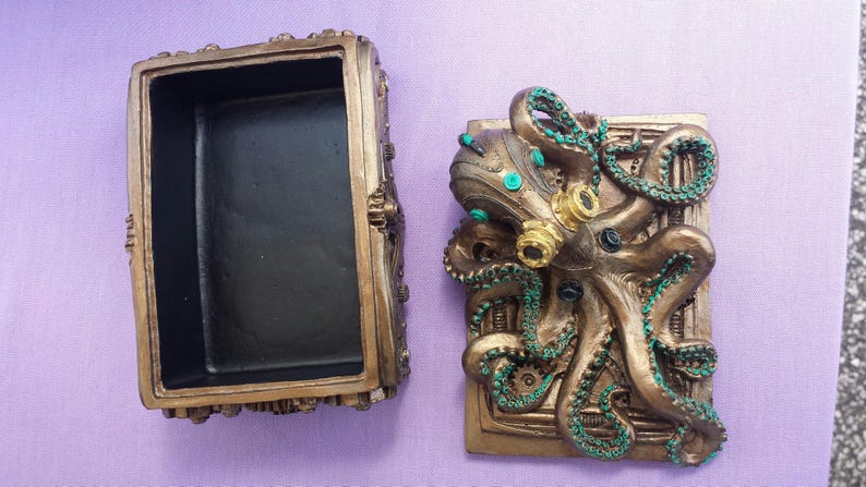 Puede incluir: Caja abierta de color bronce con un dise&ntilde;o detallado de pulpo en la tapa. El pulpo tiene detalles turquesa y dorados. La caja est&aacute; sobre una superficie morada. El interior de la caja es negro.