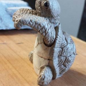 May include: A detailed, beige-colored turtle figurine with textured skin and a patterned shell. The turtle is posed with its arms outstretched, giving the impression of movement. The figurine is likely a decorative item.