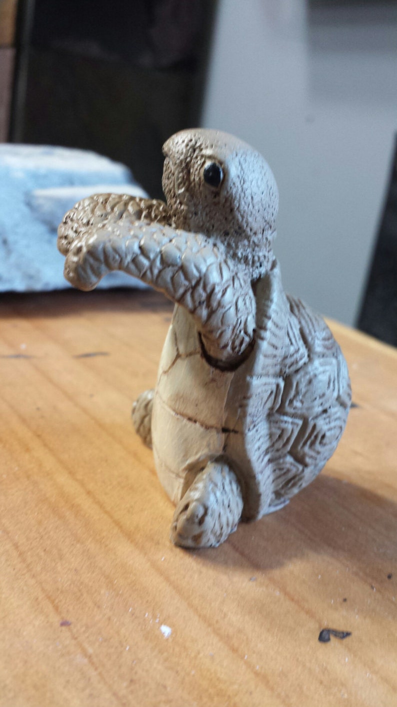 May include: A detailed, beige-colored turtle figurine with textured skin and a patterned shell. The turtle is posed with its arms outstretched, giving the impression of movement. The figurine is likely a decorative item.