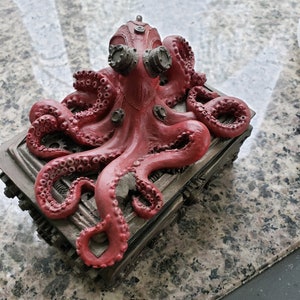 K&ouml;nnte beinhalten: Eine rote Krakenfigur mit Steampunk-Details, die auf einer Metallbox mit komplizierten Zahnr&auml;dern und Designs sitzt. Der Kraken hat gro&szlig;e, runde Augen und einen detaillierten K&ouml;rper.