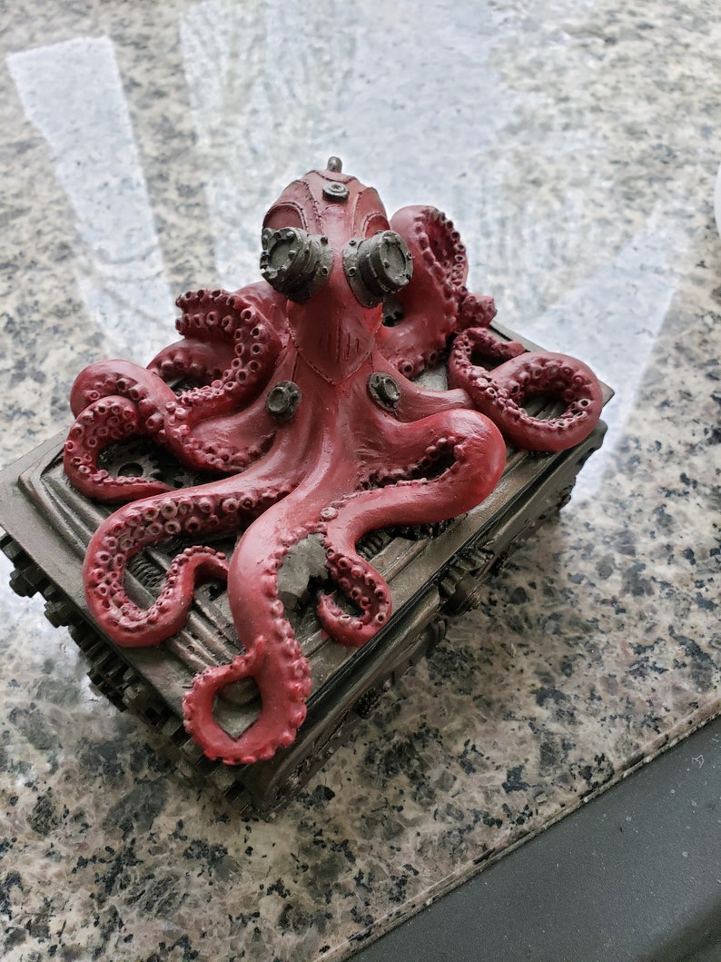 K&ouml;nnte beinhalten: Eine rote Krakenfigur mit Steampunk-Details, die auf einer Metallbox mit komplizierten Zahnr&auml;dern und Designs sitzt. Der Kraken hat gro&szlig;e, runde Augen und einen detaillierten K&ouml;rper.