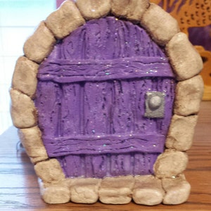 May include: A miniature purple door with a stone frame and a silver door knob. The door is decorated with glitter.