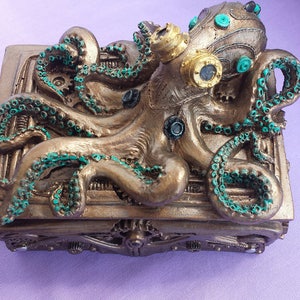 Puede incluir: Una caja de estilo steampunk con un dise&ntilde;o de pulpo. La caja es de color bronce con detalles turquesa en los tent&aacute;culos y otros elementos. El pulpo tiene gafas y elementos mec&aacute;nicos, creando una pieza art&iacute;stica &uacute;nica.