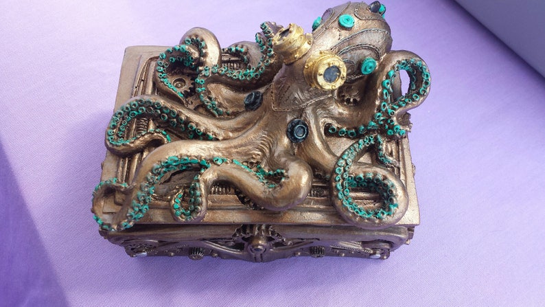 Puede incluir: Una caja de estilo steampunk con un dise&ntilde;o de pulpo. La caja es de color bronce con detalles turquesa en los tent&aacute;culos y otros elementos. El pulpo tiene gafas y elementos mec&aacute;nicos, creando una pieza art&iacute;stica &uacute;nica.