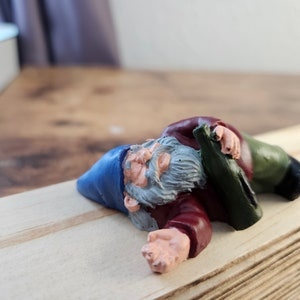 Könnte beinhalten: Eine kleine Garten-Zwergenfigur aus blauem, rotem und grünem Kunststoff liegt auf der Seite auf einer Holzoberfläche. Der Zwerg hält eine grüne Flasche in der Hand und hat einen weißen Bart.