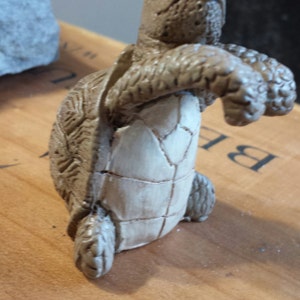 May include: A detailed figurine of a turtle, featuring a textured shell and a light-colored underbelly. The turtle is in a walking pose, with its front legs extended. The figurine is made of a light brown material.