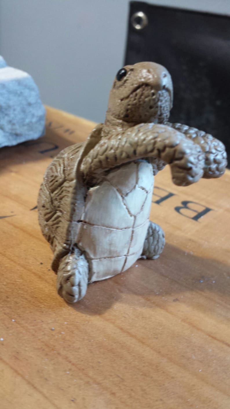 May include: A detailed figurine of a turtle, featuring a textured shell and a light-colored underbelly. The turtle is in a walking pose, with its front legs extended. The figurine is made of a light brown material.