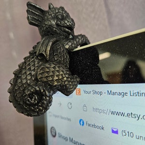 Puede incluir: Una figurita de drag&oacute;n de metal negro posada en la pantalla de un port&aacute;til. El drag&oacute;n est&aacute; mirando hacia el lado izquierdo de la imagen y tiene un cuerpo texturizado y escamoso. La pantalla del port&aacute;til muestra un sitio web con el texto "Your Shop - Manage Listings" y "https://www.etsy.com".