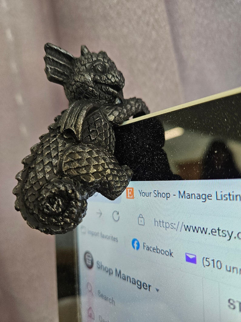 Puede incluir: Una figurita de drag&oacute;n de metal negro posada en la pantalla de un port&aacute;til. El drag&oacute;n est&aacute; mirando hacia el lado izquierdo de la imagen y tiene un cuerpo texturizado y escamoso. La pantalla del port&aacute;til muestra un sitio web con el texto "Your Shop - Manage Listings" y "https://www.etsy.com".