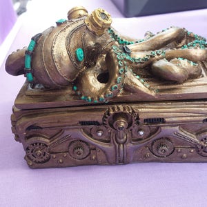 Puede incluir: Una caja de estilo steampunk con un dise&ntilde;o de pulpo. La caja es de color bronce con detalles de engranajes y acentos turquesa. El pulpo tiene tent&aacute;culos y gafas. La caja est&aacute; sobre una superficie p&uacute;rpura.