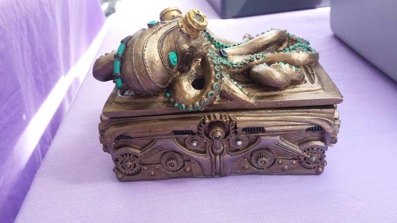 Puede incluir: Una caja de estilo steampunk con un dise&ntilde;o de pulpo. La caja es de color bronce con detalles de engranajes y acentos turquesa. El pulpo tiene tent&aacute;culos y gafas. La caja est&aacute; sobre una superficie p&uacute;rpura.