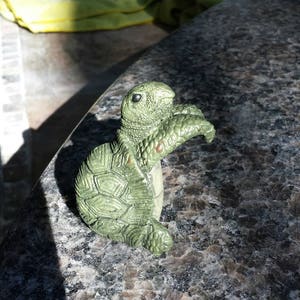 May include: A small, green turtle figurine with a textured shell and detailed features. The figurine is made of a solid material and has a light green color with darker green accents. The turtle is posed in a standing position, with its head and front flipper raised.