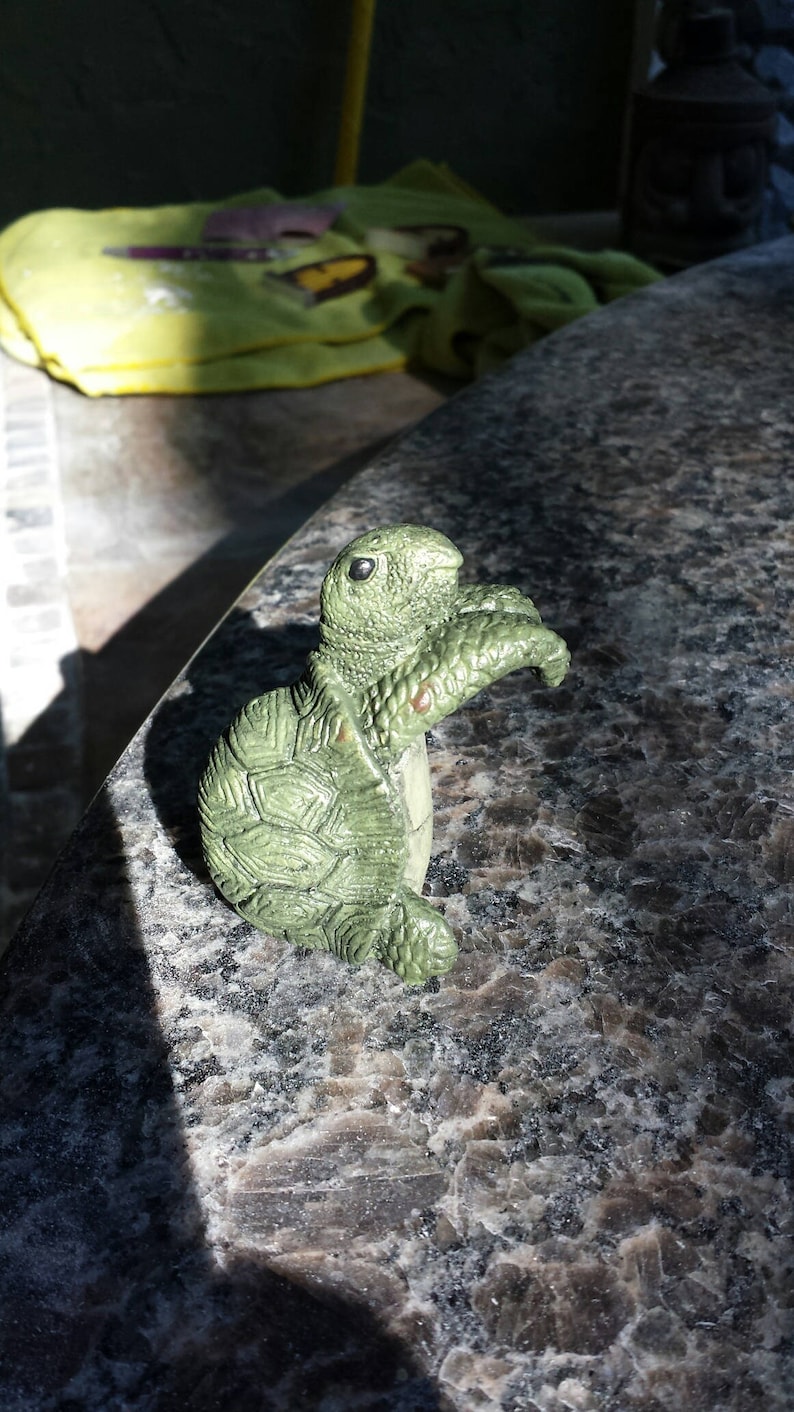 May include: A small, green turtle figurine with a textured shell and detailed features. The figurine is made of a solid material and has a light green color with darker green accents. The turtle is posed in a standing position, with its head and front flipper raised.