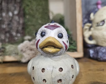 May include: A white ceramic duck with a red and black face and a spiky head. The duck has a yellow beak and black eyes. The duck is covered in small white dots.